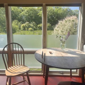 A windsor chair in light wood sits next to a round kitchen table, pulled out, as though someone is about to sit down. There is a vase of wildflowers on the table, and the greenery is echoed in the greens of the garden observed outside the window.